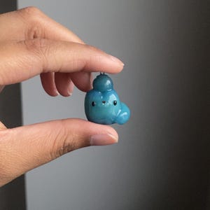 May include: A small blue and green clay figurine with a round head and a small, round body. The figurine has a smiling face with black eyes and a small, white mouth.