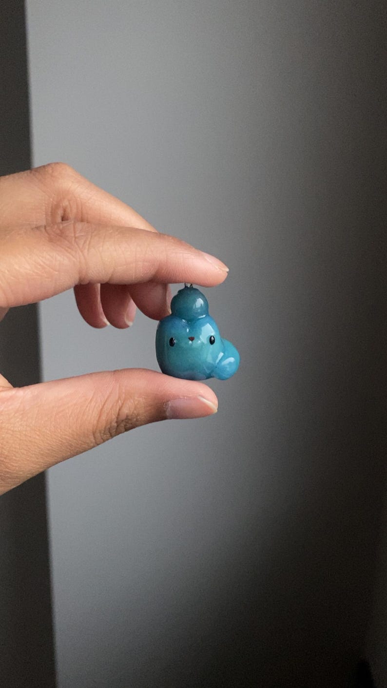May include: A small blue and green clay figurine with a round head and a small, round body. The figurine has a smiling face with black eyes and a small, white mouth.