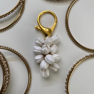 May include: A gold-colored keychain with a white flower cluster charm. Several gold bangles with various decorative elements surround the keychain. The bangles have beaded and textured designs.