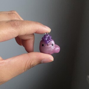May include: A small purple and pink clay charm shaped like a cartoon animal with a cluster of purple beads on top. The charm is hanging from a silver loop.