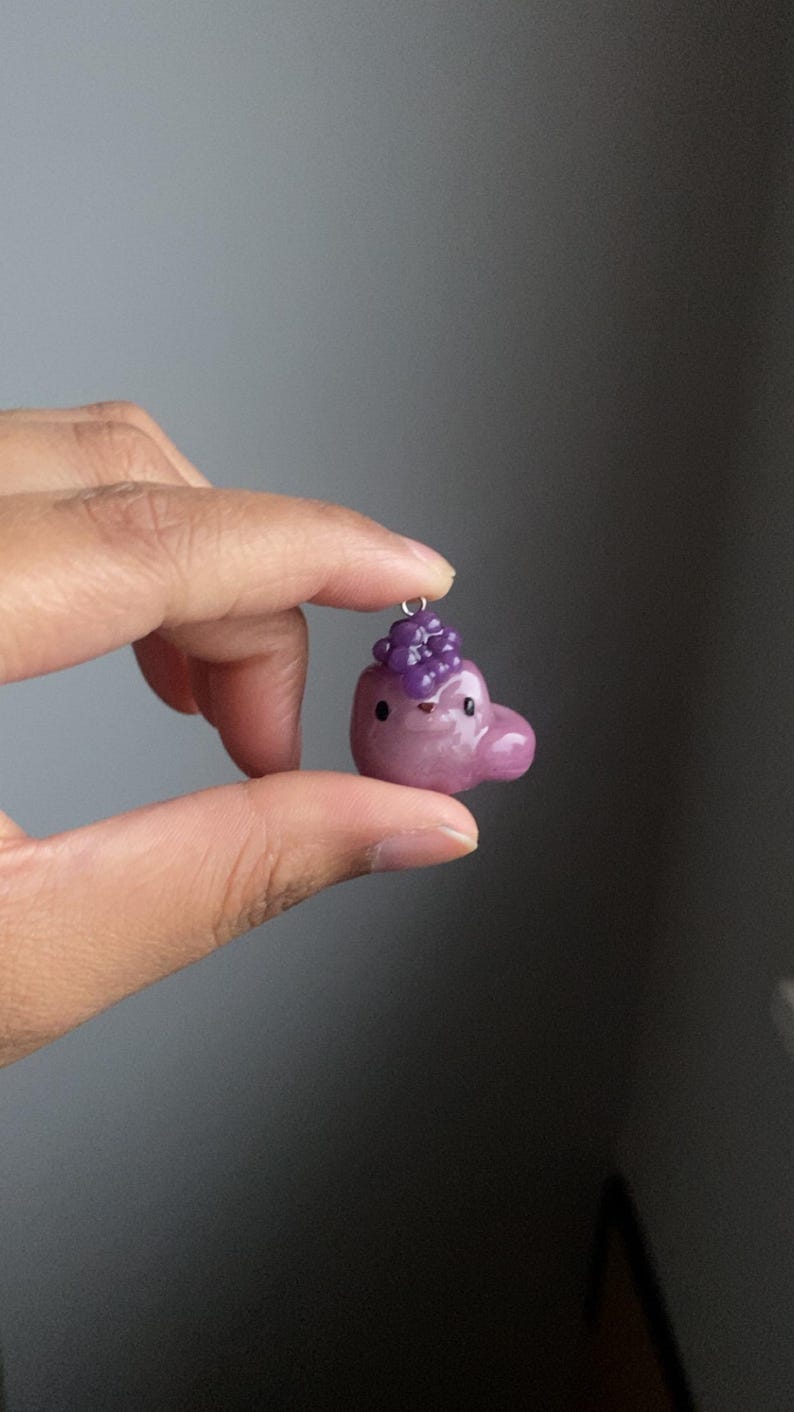 May include: A small purple and pink clay charm shaped like a cartoon animal with a cluster of purple beads on top. The charm is hanging from a silver loop.