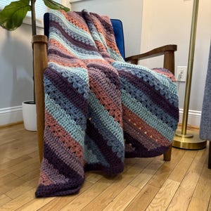 Peut inclure: Une couverture crochetée à la main drapée sur une chaise bleue. La couverture présente des rayures diagonales dans les tons de violet, de bleu et de pêche. Le motif texturé et la palette de couleurs créent une esthétique chaleureuse et invitante. Idéal pour la décoration intérieure.