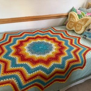 May include: A round, star-shaped crochet blanket featuring concentric rings of vibrant colors including teal, yellow, orange, and red. The blanket has a textured, repeating zigzag pattern that radiates from the center outward to the pointed edges.