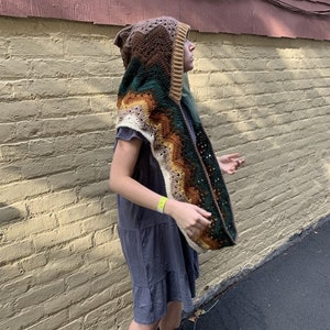 May include: A brown, green, and cream crocheted hooded scarf with a wavy pattern. The scarf is worn by a person standing in front of a brick wall.