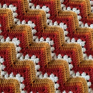 May include: Close-up of a handmade crochet blanket with a repeating pattern. The blanket features a zig-zag design in shades of red, orange, gold, and white. The crochet stitches are visible, creating a textured surface.