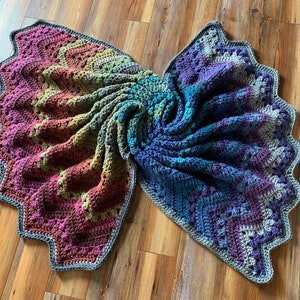 May include: A handmade crochet blanket in a butterfly wing design. The blanket features a gradient of colours, including pink, purple, green, and blue, with a grey scalloped edge. The blanket is displayed on a wooden surface.