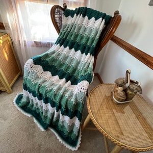 May include: A crocheted blanket featuring a repeating chevron pattern in alternating stripes of dark green, sage green, light green, and white. The blanket has a scalloped edge and is draped over a high-backed wicker chair.