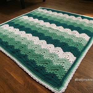 以下が含まれることがあります： 緑と白のシェブロン模様のクロシェ編み毛布。毛布は白い縁取りがされています。