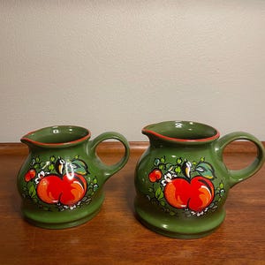 May include: Two green ceramic pitchers with a red apple design. The pitchers have a handle and a spout. The apple design includes green leaves and white flowers. The rims are trimmed in red.