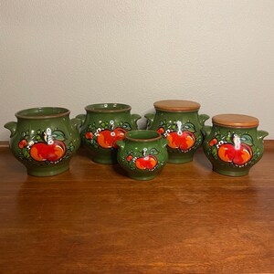 May include: Set of five green ceramic canisters with red apple illustrations and wooden lids. The canisters are arranged in a row on a wooden surface.