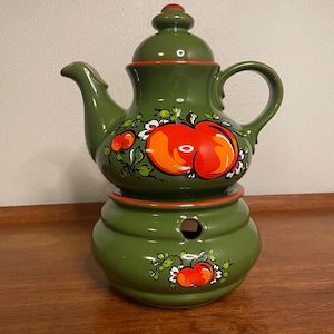 May include: A green ceramic teapot with a painted design of red apples and white flowers. The teapot has a separate base with a hole for a tea light.