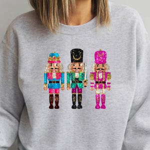 May include: A light gray sweatshirt featuring three colorful nutcracker designs. The nutcrackers have blue, green, and pink accents, with black boots and hats. The design is centered on the chest.