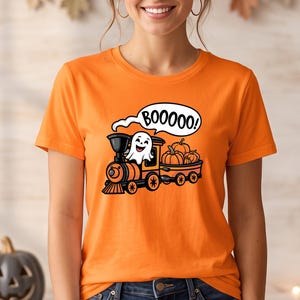 May include: An orange short-sleeve t-shirt featuring a graphic of a black and orange steam train carrying orange pumpkins. A white ghost sits in the engine cab with a speech bubble above its head containing the black text BOOOOOO.