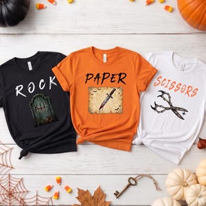 May include: Three short-sleeve t-shirts in black, orange, and white. The black shirt features the text ROCK and a tombstone. The orange shirt features the text PAPER and a knife on parchment. The white shirt features the text SCISSORS and shears.