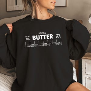 以下が含まれることがあります： 白い文字で「SALTED BUTTER GRADE AA」と書いてある黒いスウェットシャツ。大さじとカップの計量ガイド付き。テキストには正味重量が4オンス（113g）と記載されています。