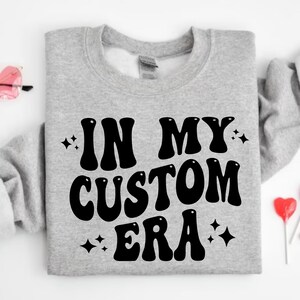 K&ouml;nnte beinhalten: Graues Sweatshirt mit dem schwarzen Schriftzug "IN MY CUSTOM ERA" in Retro-Schrift. Das Bild zeigt rosa herzf&ouml;rmige Sonnenbrillen, herzf&ouml;rmige Lollis und herzf&ouml;rmige S&uuml;&szlig;igkeiten.