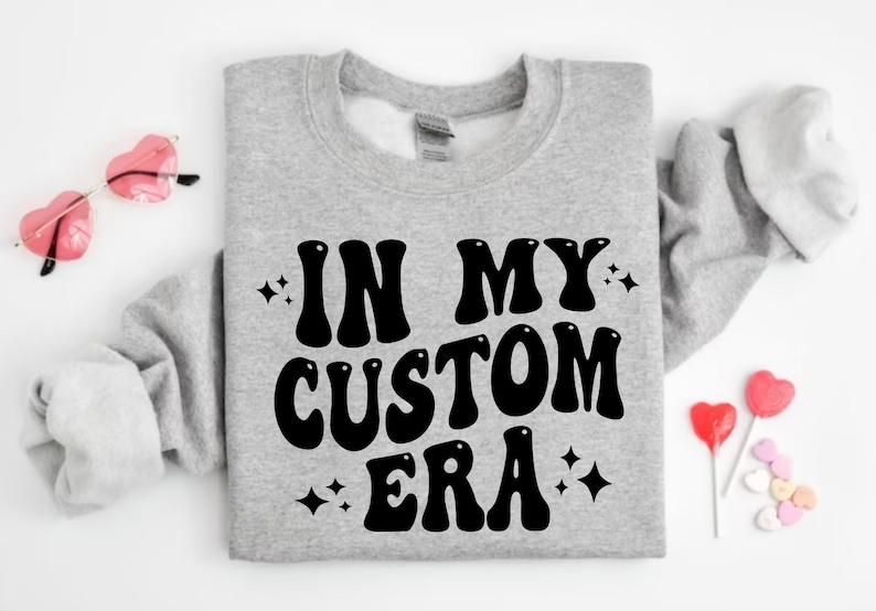 K&ouml;nnte beinhalten: Graues Sweatshirt mit dem schwarzen Schriftzug "IN MY CUSTOM ERA" in Retro-Schrift. Das Bild zeigt rosa herzf&ouml;rmige Sonnenbrillen, herzf&ouml;rmige Lollis und herzf&ouml;rmige S&uuml;&szlig;igkeiten.