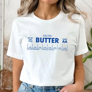 以下が含まれることがあります： バターの棒の青いグラフィックが描かれた白いTシャツ。"SALTED BUTTER GRADE AA"という文字と、大さじとカップの測定ガイドが書かれています。