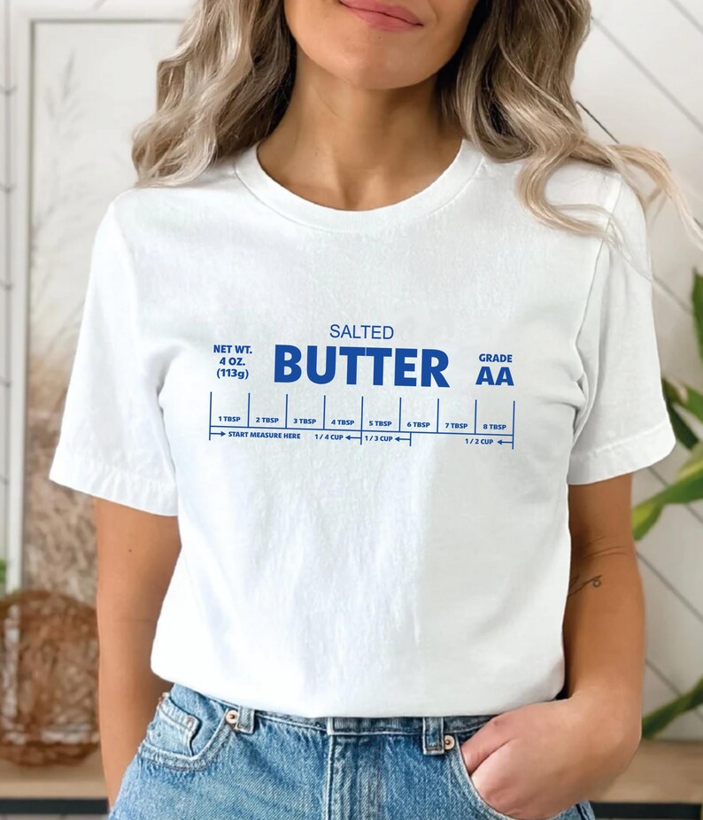 以下が含まれることがあります： バターの棒の青いグラフィックが描かれた白いTシャツ。"SALTED BUTTER GRADE AA"という文字と、大さじとカップの測定ガイドが書かれています。