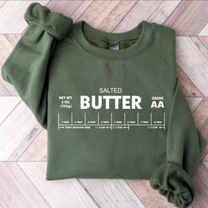以下が含まれることがあります： バターのさじとカップの測定ガイドと「SALTED BUTTER GRADE AA」のテキストが書かれた緑色のスウェットシャツ。「NET WT. 4 OZ. (113g)」のテキストも含まれています。