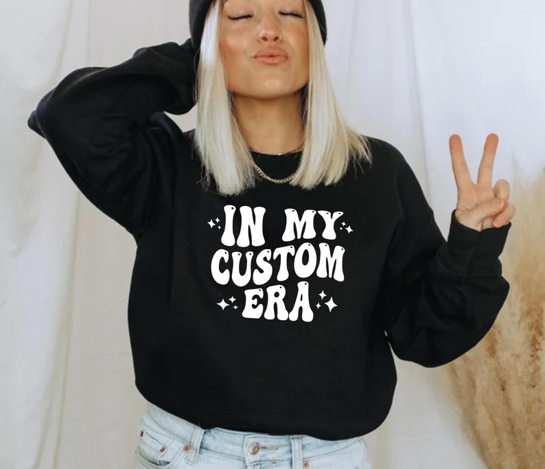 K&ouml;nnte beinhalten: Schwarzes Sweatshirt mit dem wei&szlig;en Text "IN MY CUSTOM ERA" in Retro-Schrift. Das Sweatshirt hat einen Rundhalsausschnitt und lange &Auml;rmel. Kleine Sternchen umgeben den Text.