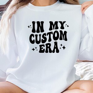 K&ouml;nnte beinhalten: Wei&szlig;es Sweatshirt mit dem Text "IN MY CUSTOM ERA" in schwarzer, retroartiger Schrift. Das Design enth&auml;lt Sterne. Das Sweatshirt ist ein l&auml;ssiges Kleidungsst&uuml;ck mit langen &Auml;rmeln.