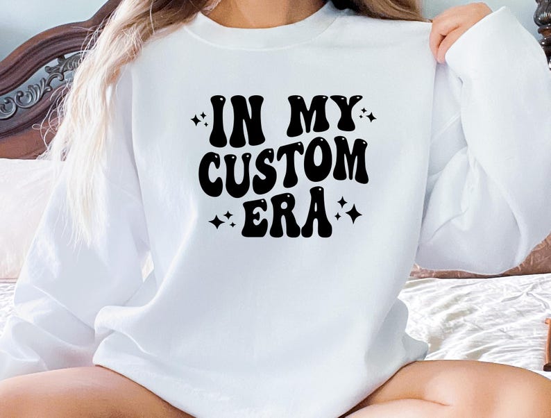 K&ouml;nnte beinhalten: Wei&szlig;es Sweatshirt mit dem Text "IN MY CUSTOM ERA" in schwarzer, retroartiger Schrift. Das Design enth&auml;lt Sterne. Das Sweatshirt ist ein l&auml;ssiges Kleidungsst&uuml;ck mit langen &Auml;rmeln.