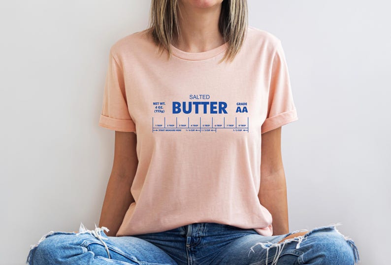 以下が含まれることがあります： 青いグラフィックが印刷されたピンクのTシャツ。グラフィックには「SALTED BUTTER GRADE AA」と、その下にスケールが描かれています。このグラフィックは、バタースティックのラベルのパロディーです。