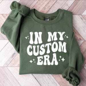 K&ouml;nnte beinhalten: Ein olivgr&uuml;nes Sweatshirt mit dem Schriftzug "IN MY CUSTOM ERA" in gro&szlig;en, wei&szlig;en Buchstaben im Retro-Stil. Das Design umfasst kleine Sternchen-Akzente. Das Sweatshirt ist aus weichem Material.
