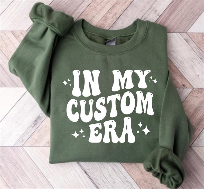 K&ouml;nnte beinhalten: Ein olivgr&uuml;nes Sweatshirt mit dem Schriftzug "IN MY CUSTOM ERA" in gro&szlig;en, wei&szlig;en Buchstaben im Retro-Stil. Das Design umfasst kleine Sternchen-Akzente. Das Sweatshirt ist aus weichem Material.
