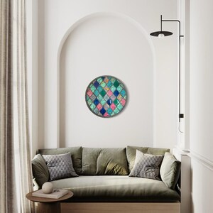 May include: A round wall art piece with a geometric pattern of teal, blue, pink, and white squares. The art is hanging on a white wall in a living room setting.
