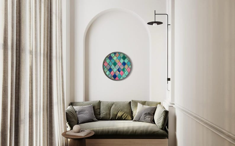 May include: A round wall art piece with a geometric pattern of teal, blue, pink, and white squares. The art is hanging on a white wall in a living room setting.