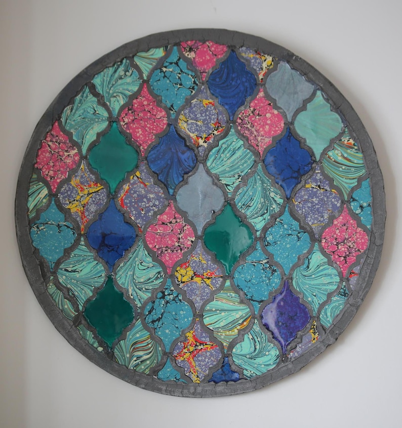 May include: A round, decorative wall hanging with a mosaic design. The mosaic is made of small, colorful tiles in shades of blue, green, pink, and purple. The tiles are arranged in a geometric pattern, creating a visually appealing and intricate design.