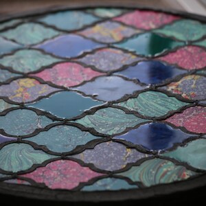 May include: A circular table top with a mosaic design. The mosaic is made of small, irregularly shaped pieces of glass in shades of blue, green, pink, and white. The pieces are arranged in a repeating pattern of interlocking shapes.