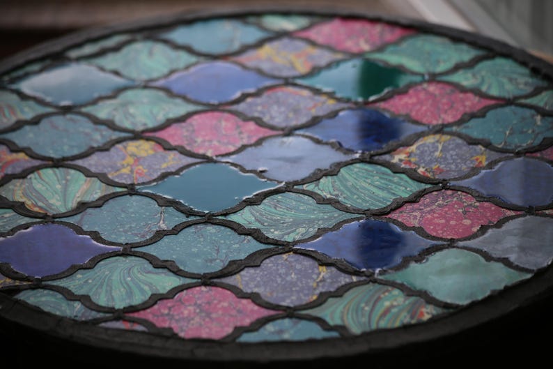 May include: A circular table top with a mosaic design. The mosaic is made of small, irregularly shaped pieces of glass in shades of blue, green, pink, and white. The pieces are arranged in a repeating pattern of interlocking shapes.
