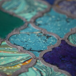 May include: A close-up of a decorative tile with a geometric pattern. The tile is made of a light blue, green, and purple marbled material. The tile has a gray border.