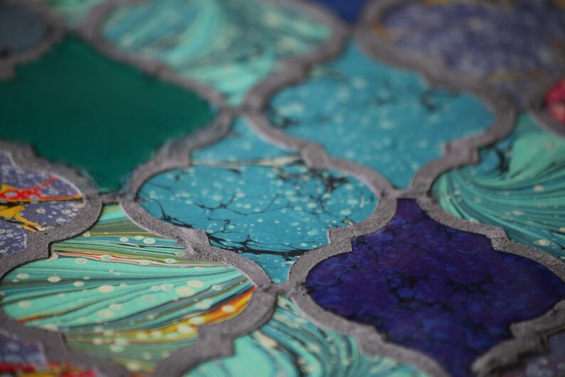 May include: A close-up of a decorative tile with a geometric pattern. The tile is made of a light blue, green, and purple marbled material. The tile has a gray border.