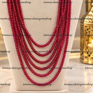 May include: A multi-strand necklace featuring six cascading layers of small, round, deep red beads. The strands are gathered at the top with a red fabric-wrapped cord. The text "Immaculategemsshop" is repeated across the image.