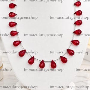 May include: A necklace featuring a series of faceted, teardrop-shaped red gemstones strung along a thin silver-toned wire. The stones are arranged symmetrically, with the largest drops positioned at the centre and smaller ones toward the ends.