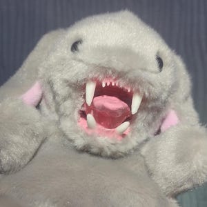 以下が含まれることがあります： 鋭い白い歯とピンクの舌を見せる、大きく開いた口を持つウサギの形をした灰色のぬいぐるみ。ウサギはピンク色の内側の耳をしており、直立しています。