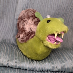 Puede incluir: Un peluche de caracol monstruo verde con una concha marrón y difusa. El caracol tiene una boca muy abierta con dientes afilados y una lengua púrpura. El juguete tiene pequeñas antenas verdes y una pequeña gema transparente en la cabeza.