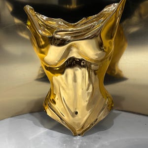 Op de afbeelding: Een gepolijst goudkleurig beeldhouwwerk in de vorm van een menselijke torso. Het object heeft een sterk reflecterend, metalen oppervlak met gladde, vloeiende contouren die de anatomie van de borst, ribbenkast en buik tot aan de taille nabootsen.