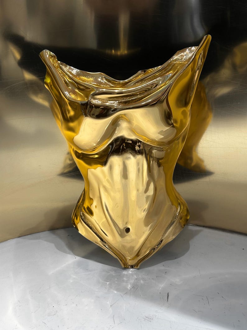 Op de afbeelding: Een gepolijst goudkleurig beeldhouwwerk in de vorm van een menselijke torso. Het object heeft een sterk reflecterend, metalen oppervlak met gladde, vloeiende contouren die de anatomie van de borst, ribbenkast en buik tot aan de taille nabootsen.