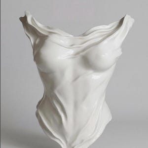 Op de afbeelding: Een wit keramisch beeldhouwwerk dat een menselijke torso voorstelt met een gedrapeerde, stofachtige textuur. Het object heeft een gladde, glanzende afwerking met diepe plooien en contouren die het uiterlijk van vloeiende stof nabootsen.