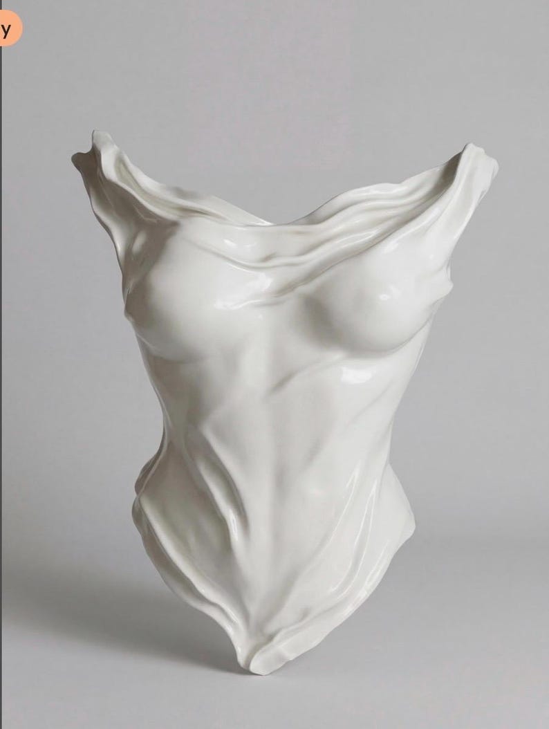 Op de afbeelding: Een wit keramisch beeldhouwwerk dat een menselijke torso voorstelt met een gedrapeerde, stofachtige textuur. Het object heeft een gladde, glanzende afwerking met diepe plooien en contouren die het uiterlijk van vloeiende stof nabootsen.