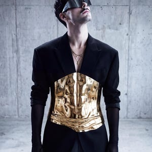 May include: A person wearing a black blazer with a gold-colored, textured corset. The person is also wearing dark sunglasses and black gloves. The background is a concrete wall.