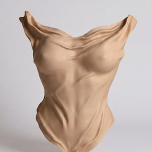 Op de afbeelding: Een beige wandsculptuur die een menselijke romp voorstelt. Het object heeft een gladde, matte afwerking met realistische, stofachtige plooien die de borst en buikstreek accentueren en een gevoel van diepte en beweging cre&euml;ren.