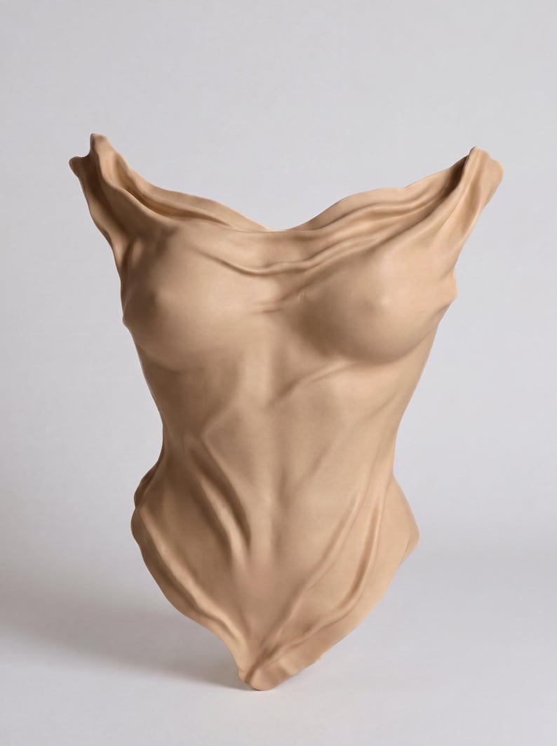 Op de afbeelding: Een beige wandsculptuur die een menselijke romp voorstelt. Het object heeft een gladde, matte afwerking met realistische, stofachtige plooien die de borst en buikstreek accentueren en een gevoel van diepte en beweging cre&euml;ren.