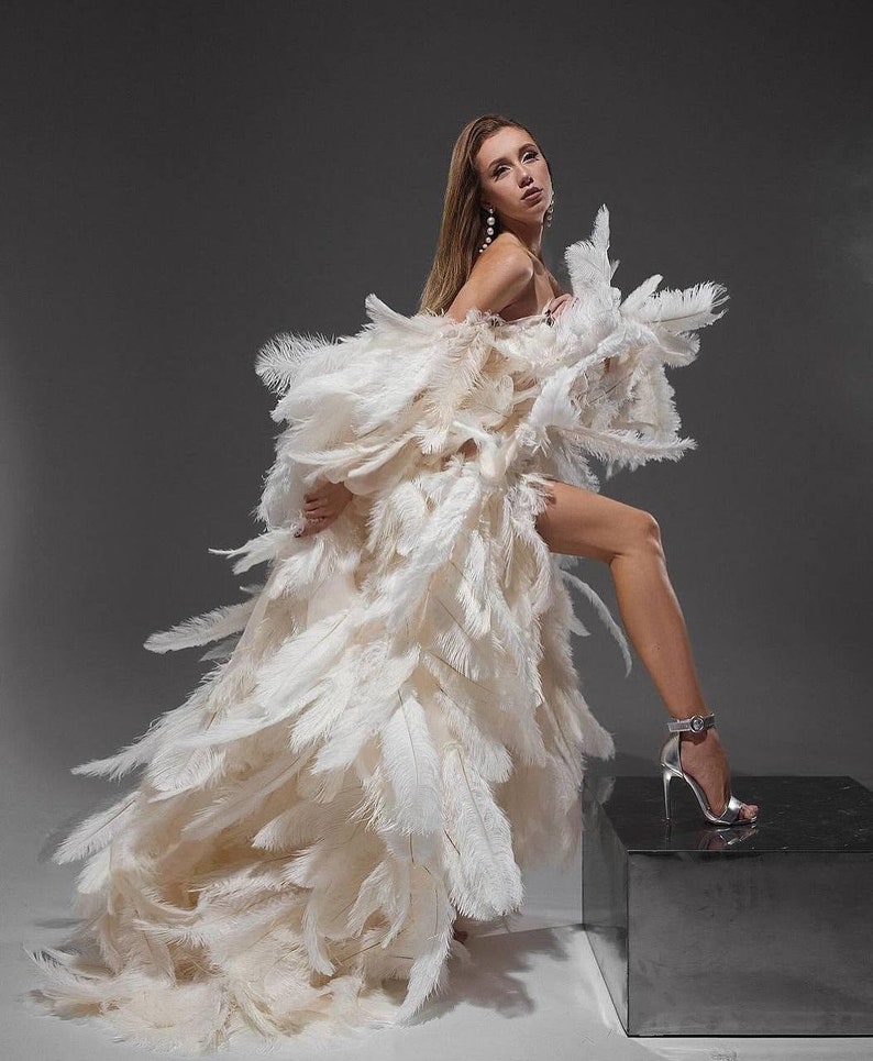 K&ouml;nnte beinhalten: Eine Frau in einem wei&szlig;en Federkleid mit langem Schlepp. Sie steht auf einer silbernen Plattform, ein Bein ist angehoben, und sie tr&auml;gt silberne High Heels.