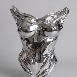 Op de afbeelding: Een sculptuur van een torso in gepolijst zilver met een sterk reflecterend, vloeibaar metalen oppervlak met vloeiende, golvende contouren die de vorm van een menselijk bovenlichaam nabootsen, inclusief gedefinieerde borst- en buikgebieden.
